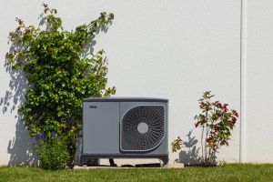 Wärmepumpe an einer weißen Hauswand mit Bepflanzung.